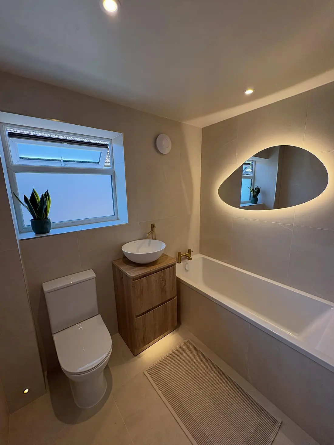 Beige bathroom in Kennington with a bath, an oak vanity with a round basin and a backlit pebble-shaped mirror