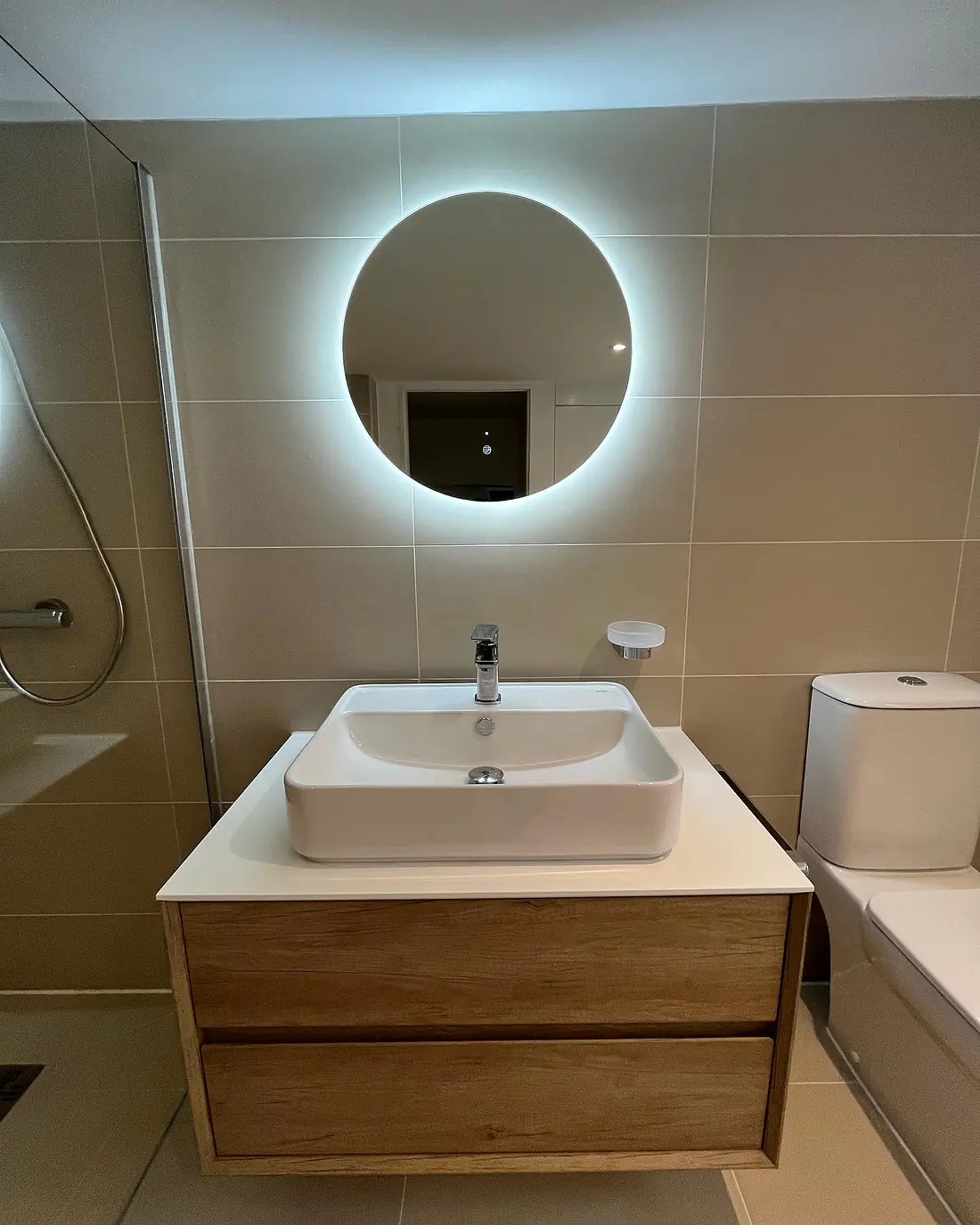 Large round backlit mirror above a white basin on a wall-hung oak vanity in a wet room