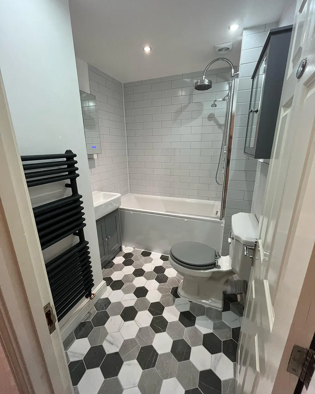 Bathroom with hexagon floor tiles, a grey vanity unit, a black radiator and a bath with a glass screen